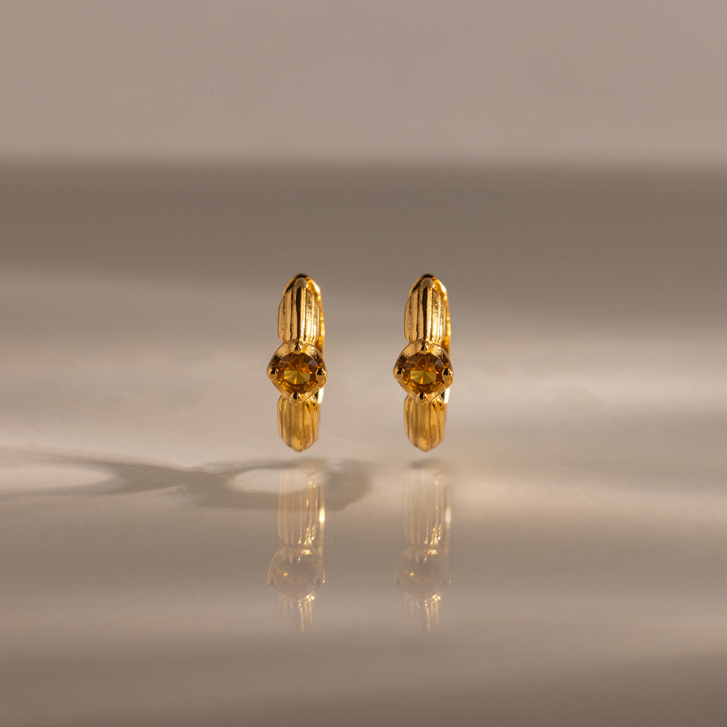 A pair of Ribbed Topaz Huggies gold hoop earrings with yellow gemstones displayed on a reflective, neutral surface.