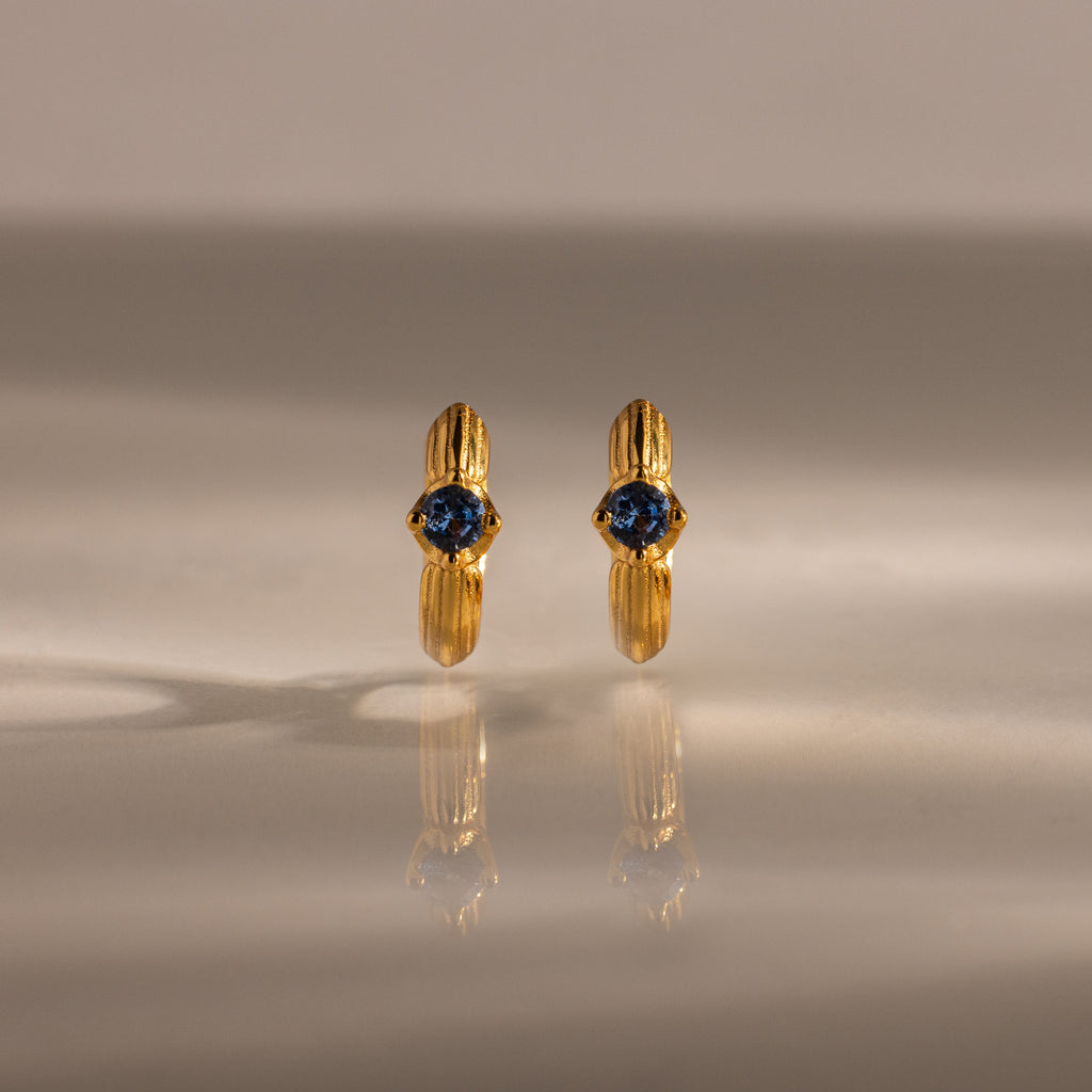 The Ribbed Blue Zircon Huggies, gold hoop earrings with blue zircon centers, are shown on a reflective surface, making this personalized jewelry sparkle.