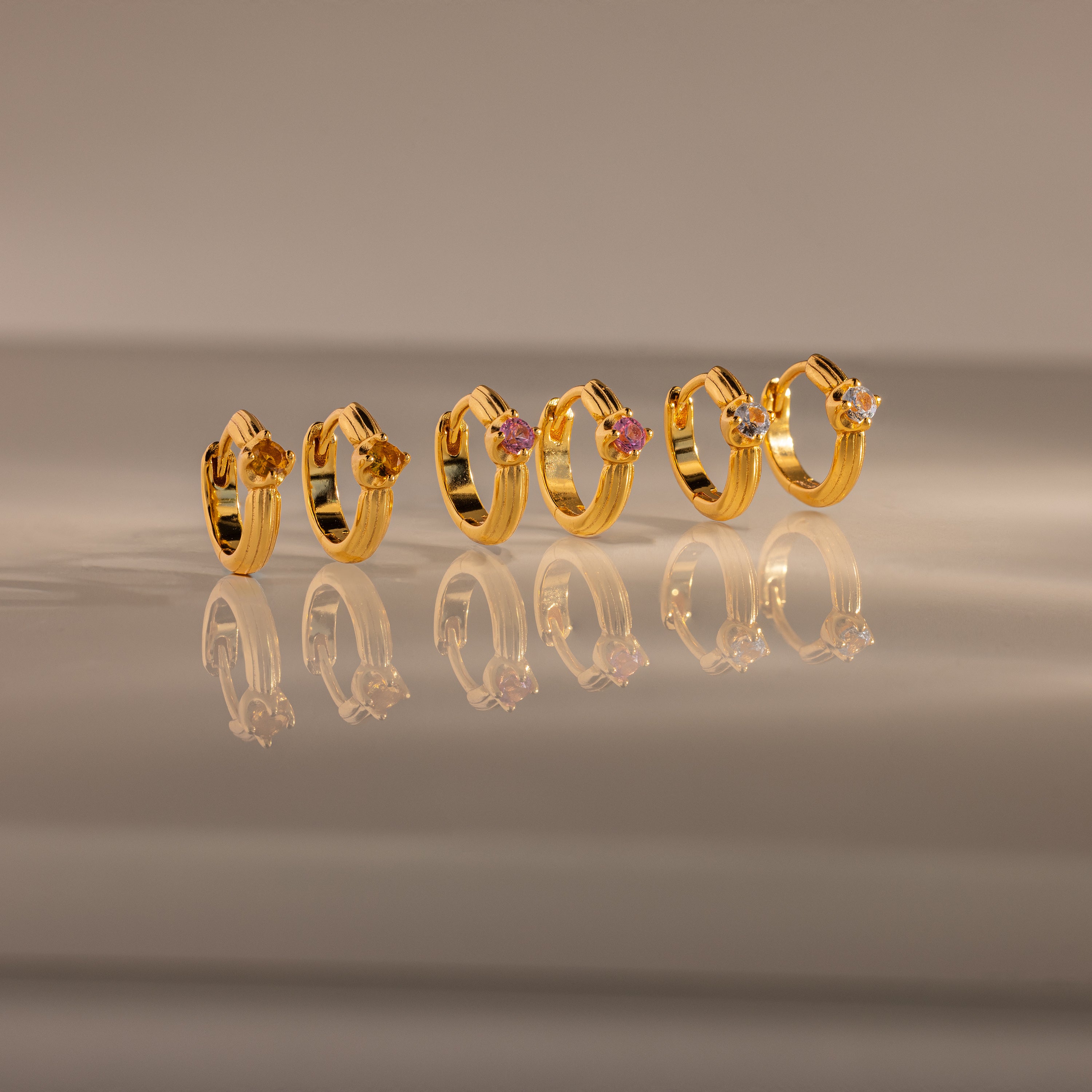 Five gold rings with colored gemstones, including Ribbed Emerald Huggies, are displayed in a row and reflected on a glossy surface—a perfect meaningful gift.
