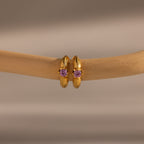 Ribbed Alexandrite Huggies—gold hoop earrings featuring purple gemstones—displayed on a light brown branch with a beige background.