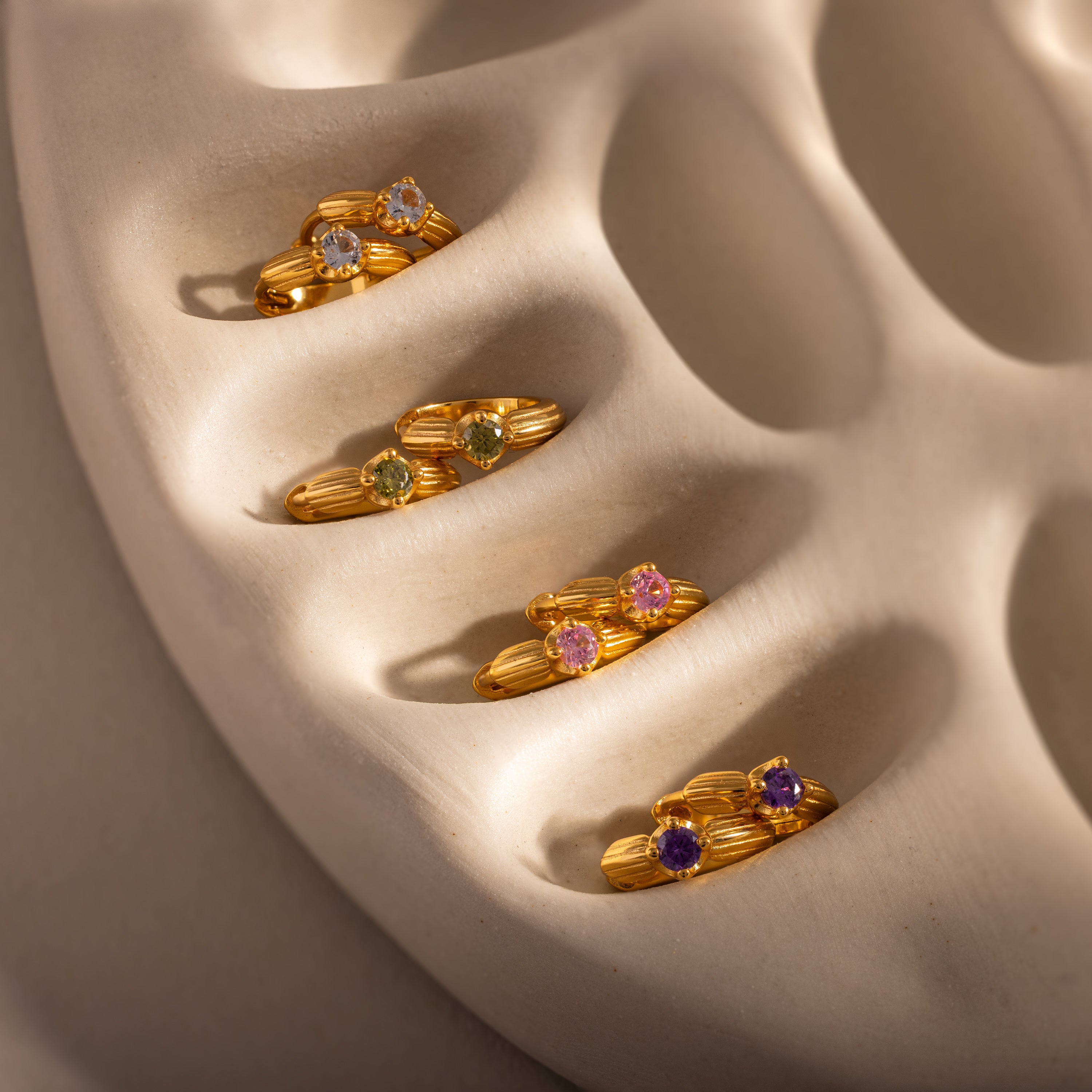 Five gold hoop earrings with emerald gemstones, inspired by Ribbed Emerald Huggies, displayed on a textured beige surface with soft shadows—a meaningful gift for any occasion.