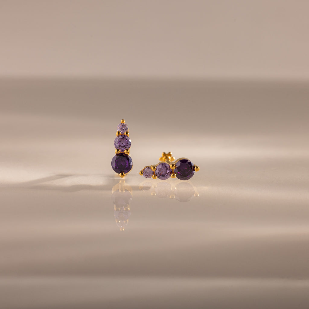 Ombre Amethyst Flatback Studs feature gold earrings with three stacked purple gemstones, shown on a smooth, reflective surface—ideal as a personalized gift or stylish flatback studs.