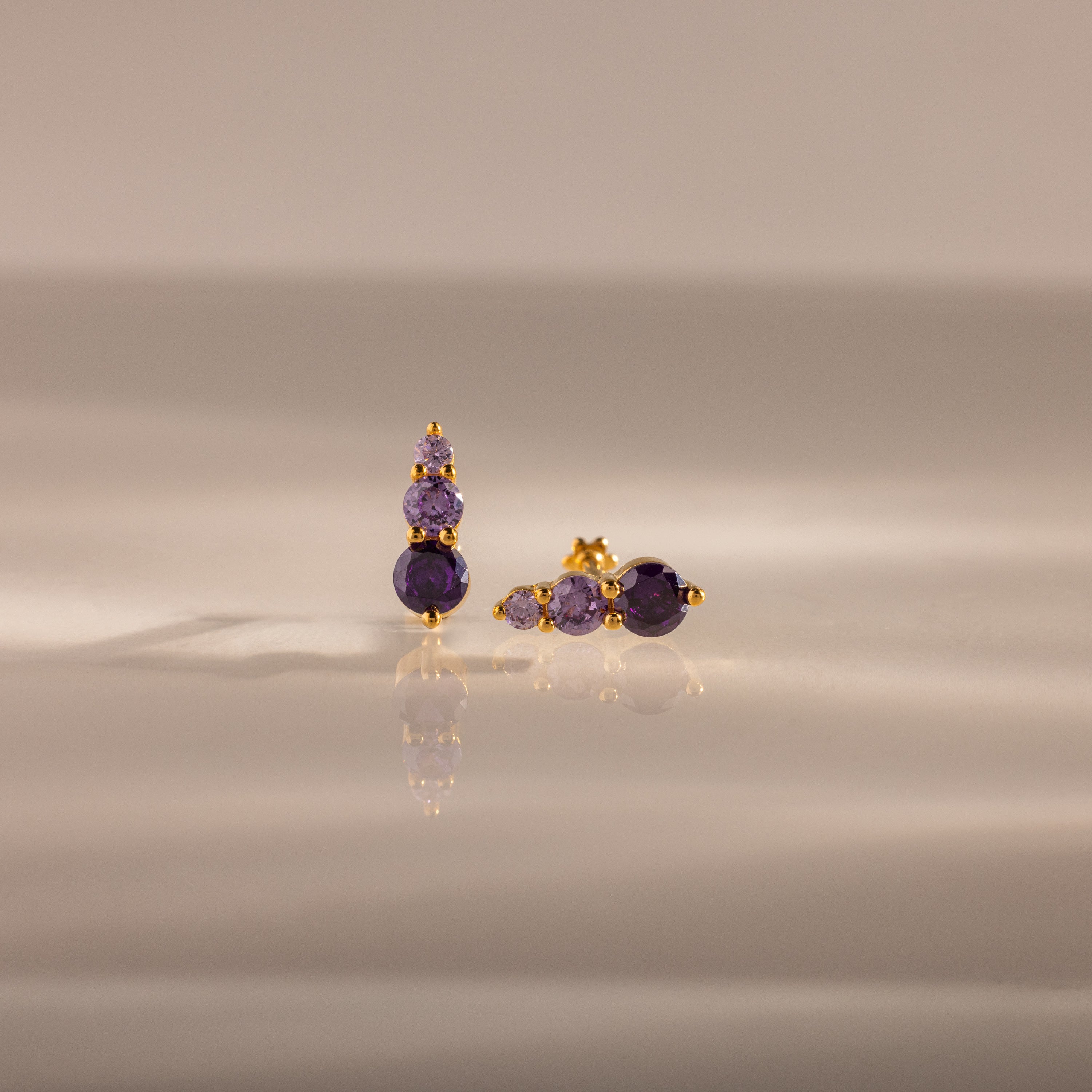 Ombre Amethyst Flatback Studs feature gold earrings with three stacked purple gemstones, shown on a smooth, reflective surface—ideal as a personalized gift or stylish flatback studs.