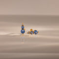 The Ombre Aquamarine Flatback Studs, each with three blue gemstones, are shown on a reflective neutral surface—these earrings make a stunning personalized gift.