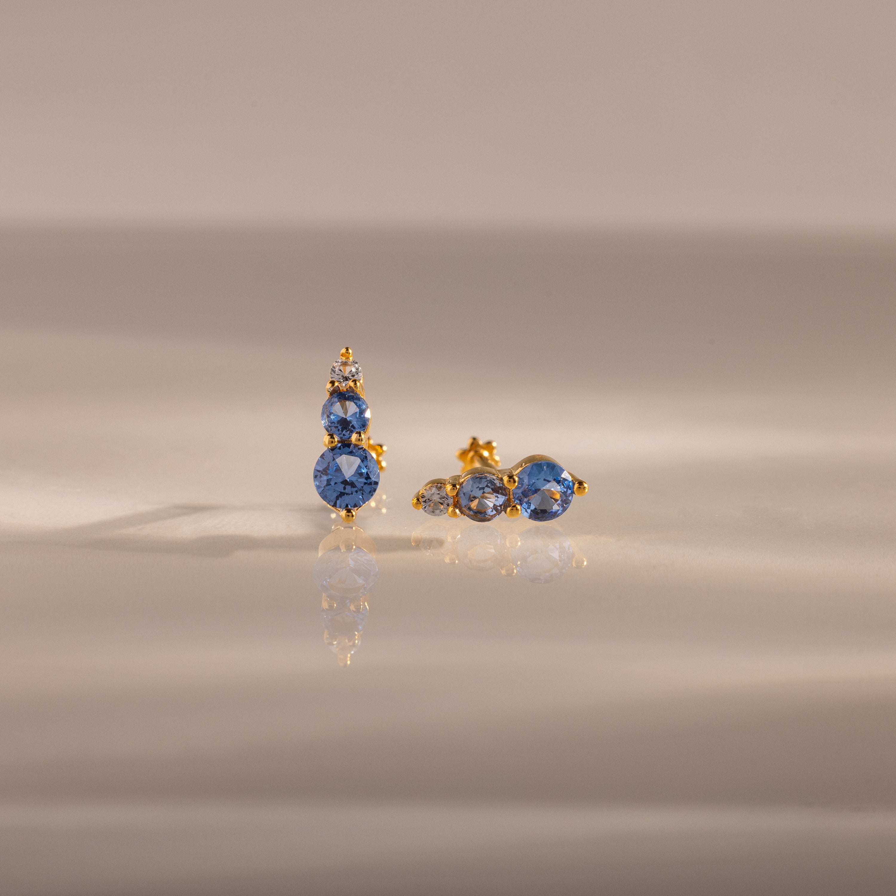 The Ombre Aquamarine Flatback Studs, each with three blue gemstones, are shown on a reflective neutral surface—these earrings make a stunning personalized gift.