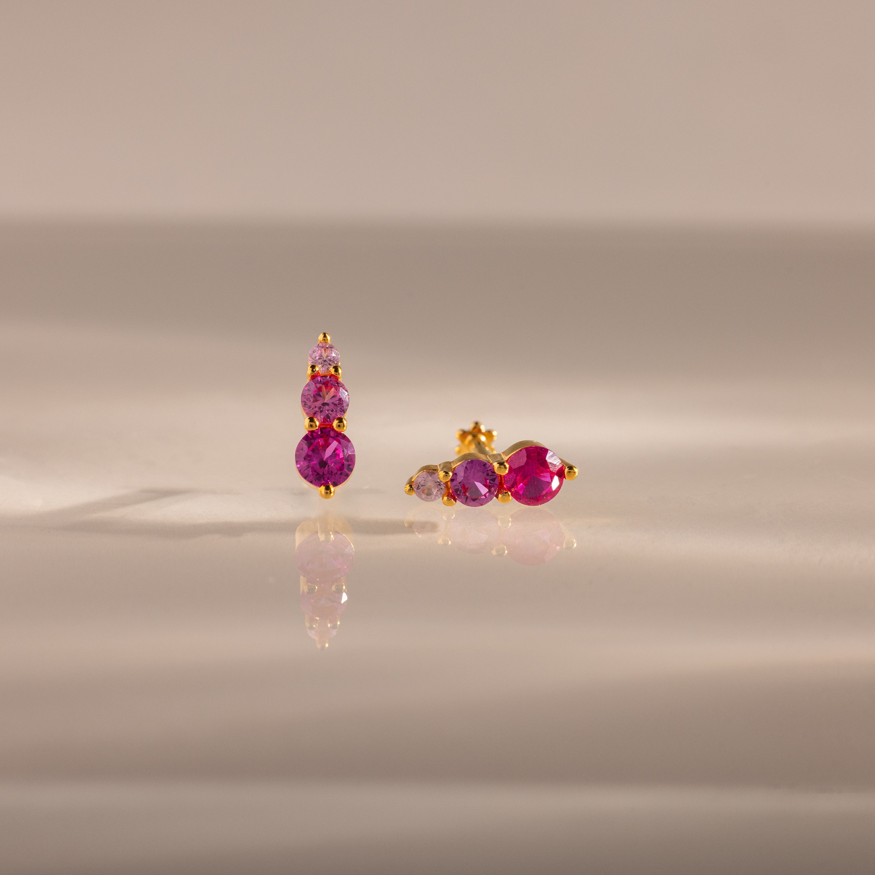 Ombre Birthstone Flatback Studs in Ruby (July) with three round pink gemstones rest on a reflective beige surface—ideal as personalized birthstone jewelry.
