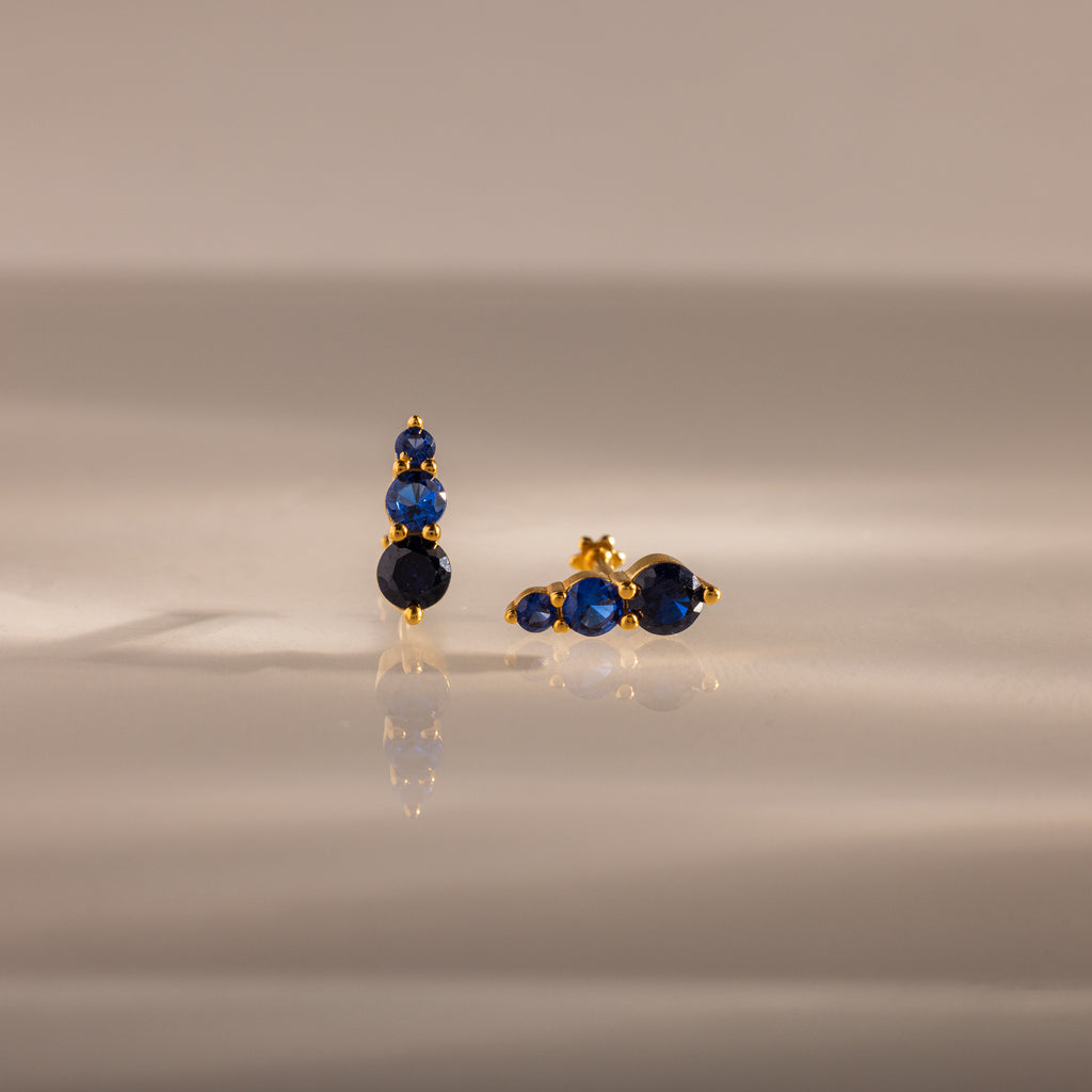 Ombre Sapphire Flatback Studs featuring three blue gemstones on a smooth, reflective beige surface—elegant, personalized jewelry for any occasion.