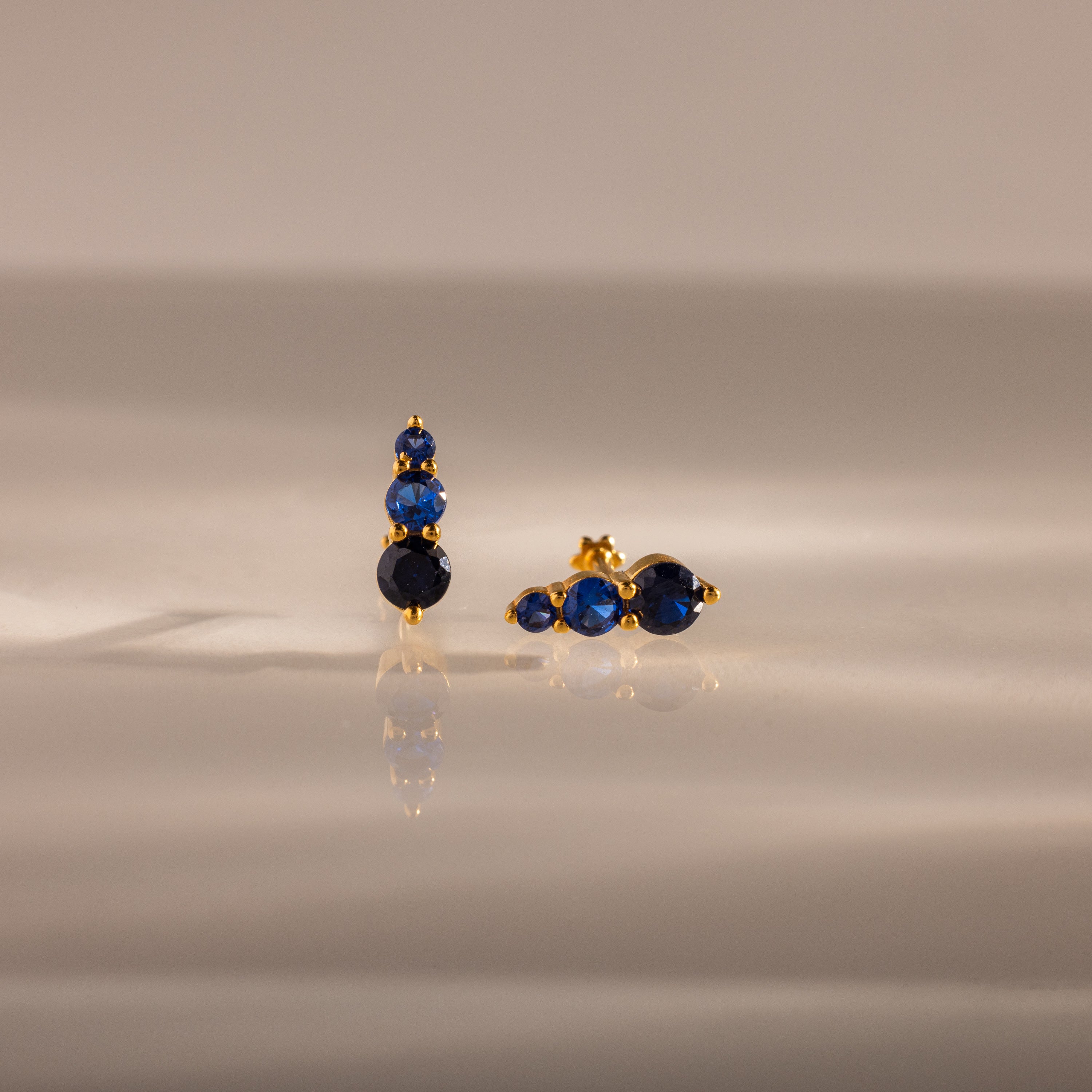 Ombre Sapphire Flatback Studs featuring three blue gemstones on a smooth, reflective beige surface—elegant, personalized jewelry for any occasion.