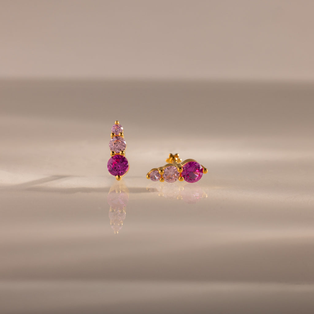 Ombre Pink Tourmaline Flatback Studs feature gold settings with three pink gemstones—one earring vertical, one horizontal—for a striking ombre birthstone effect on a reflective surface.