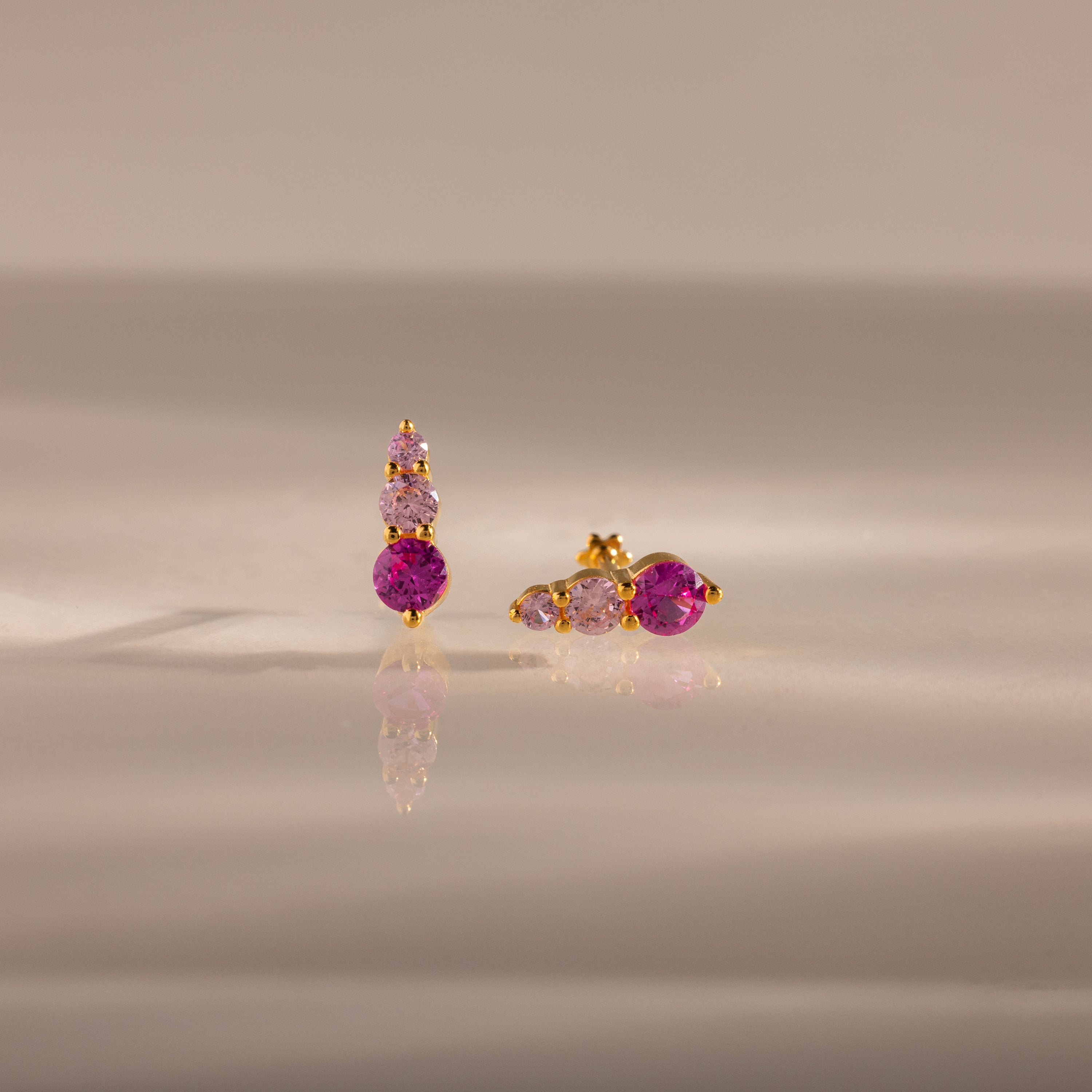 Ombre Pink Tourmaline Flatback Studs feature gold settings with three pink gemstones—one earring vertical, one horizontal—for a striking ombre birthstone effect on a reflective surface.
