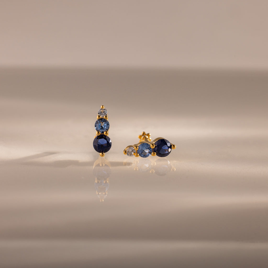 Ombre Blue Zircon Flatback Studs feature three blue zircon gems and a small clear stone on a gold setting—an elegant, personalized gift idea, beautifully displayed on a reflective surface.