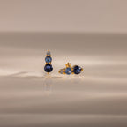 Ombre Blue Zircon Flatback Studs feature three blue zircon gems and a small clear stone on a gold setting—an elegant, personalized gift idea, beautifully displayed on a reflective surface.