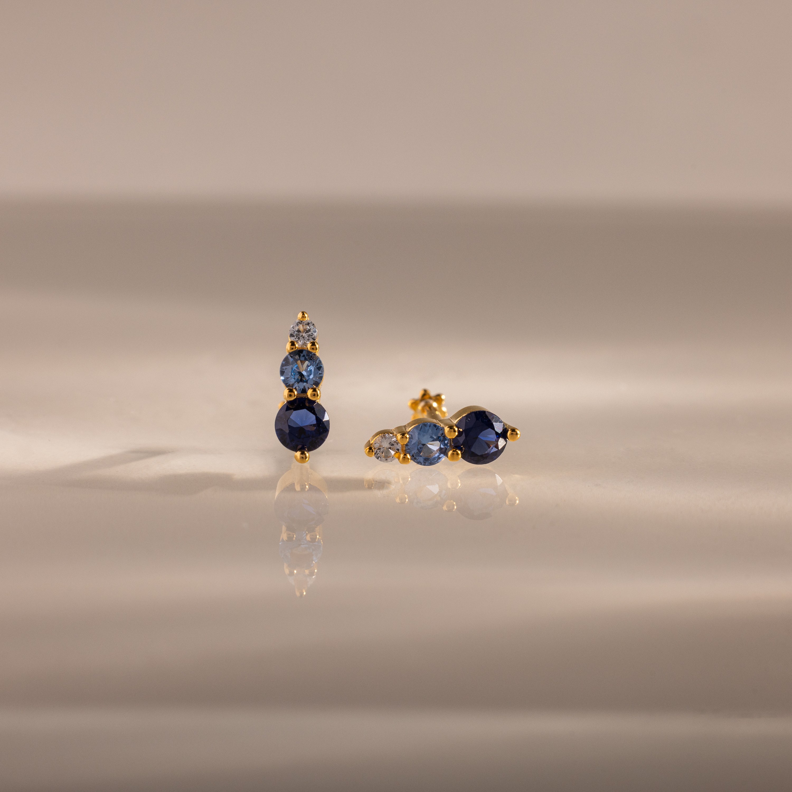 Ombre Blue Zircon Flatback Studs feature three blue zircon gems and a small clear stone on a gold setting—an elegant, personalized gift idea, beautifully displayed on a reflective surface.