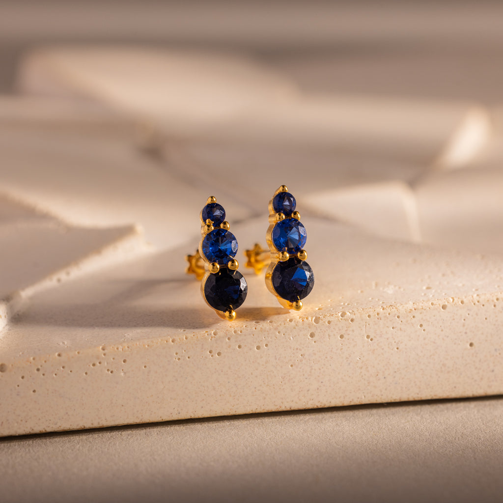 Ombre Sapphire Flatback Studs with three round blue gemstones are displayed on a textured cream surface.