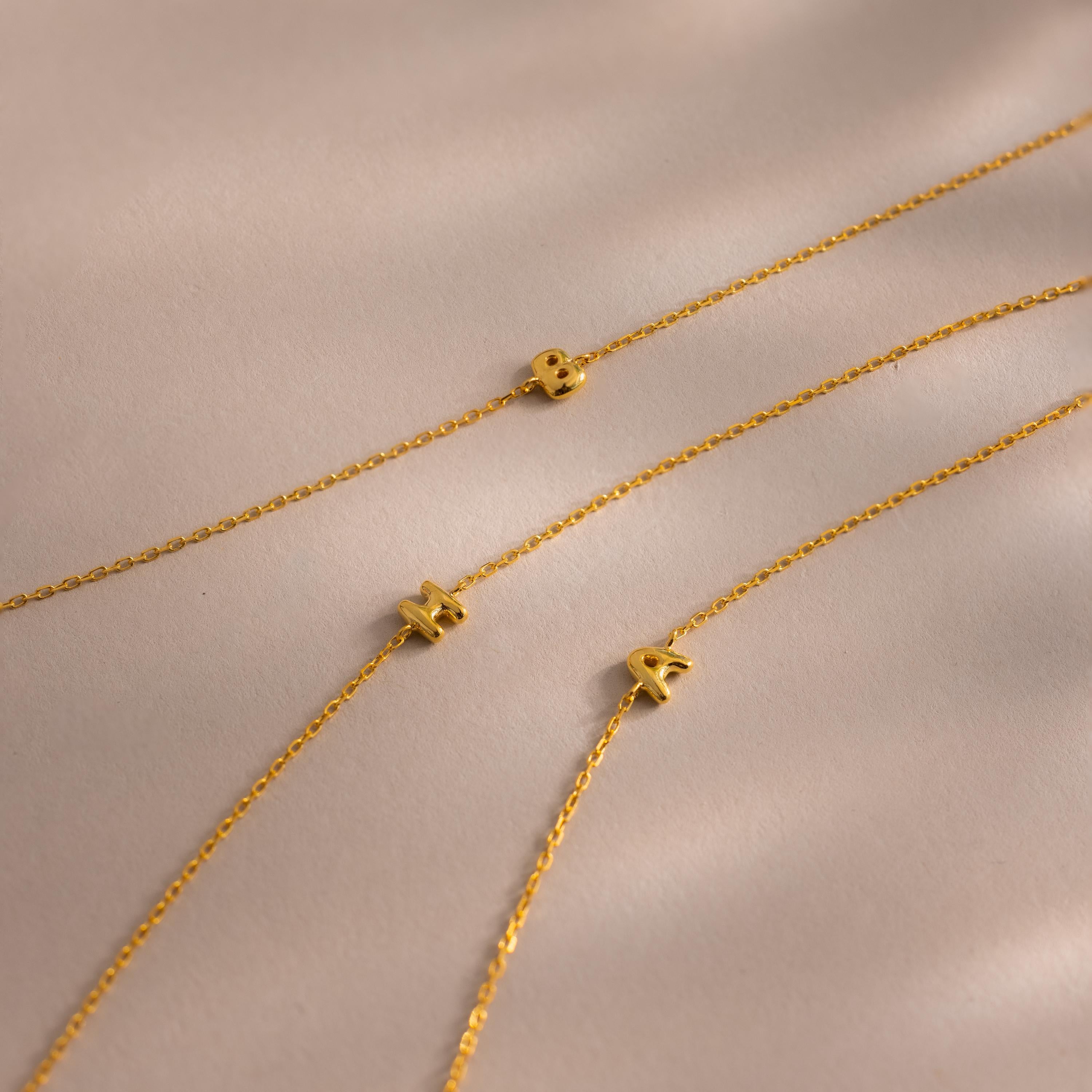 Three gold Bubble Initial Bracelets display different letters, B, M, and A, arranged on a neutral-toned background.