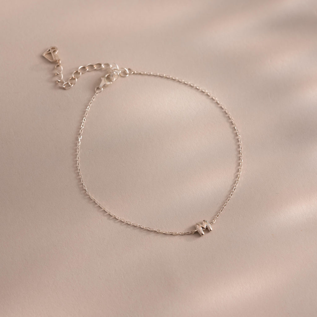Close up of a dainty silver bubble letter M bracelet against a neutral background.