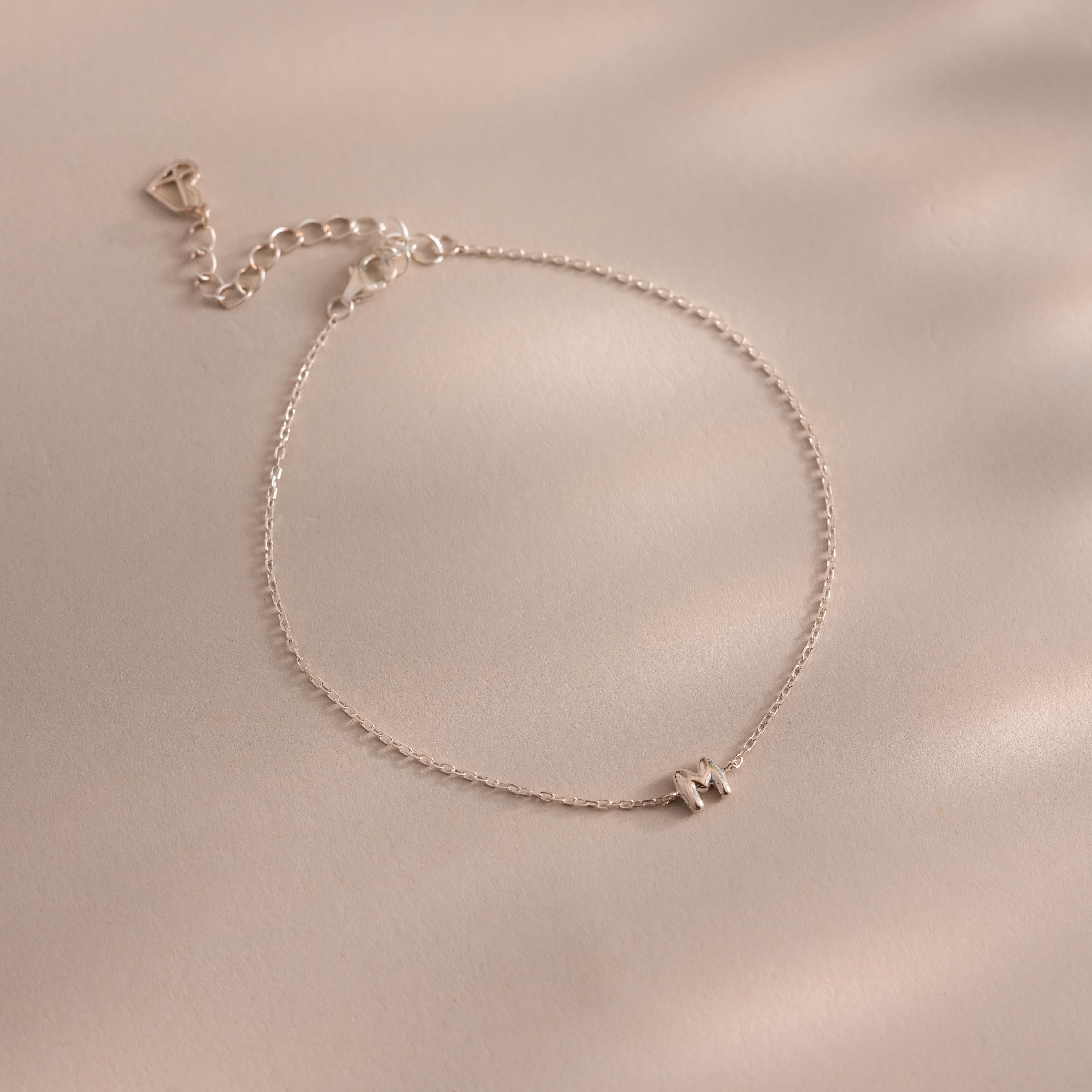 Close up of a dainty silver bubble letter M bracelet against a neutral background.