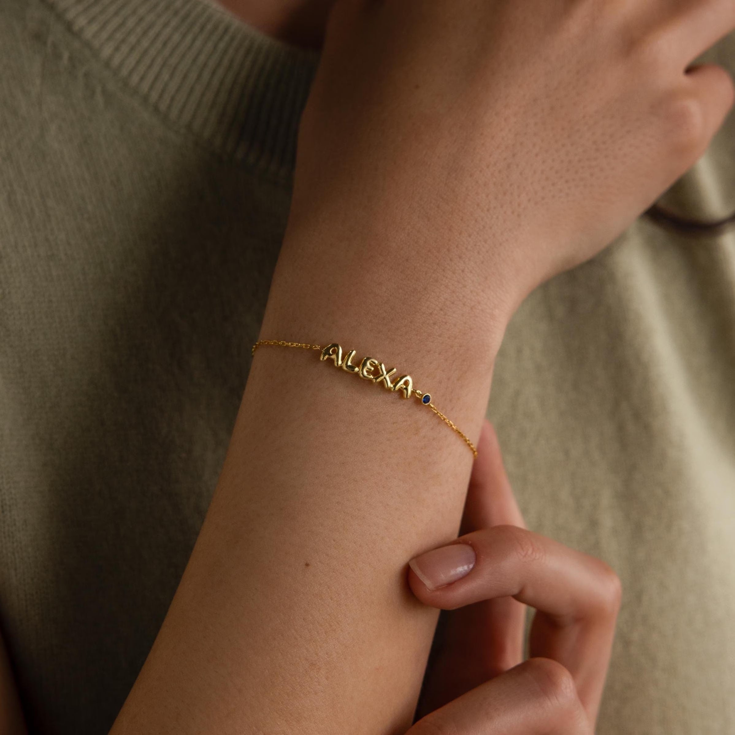 A gold bracelet spelling “ALEXA” in bold bubble-style letters is shown on a woman’s wrist, with a small sapphire birthstone charm accenting the end of the name. The bubble letters are soft, dainty with a smooth finish on a cable chain.