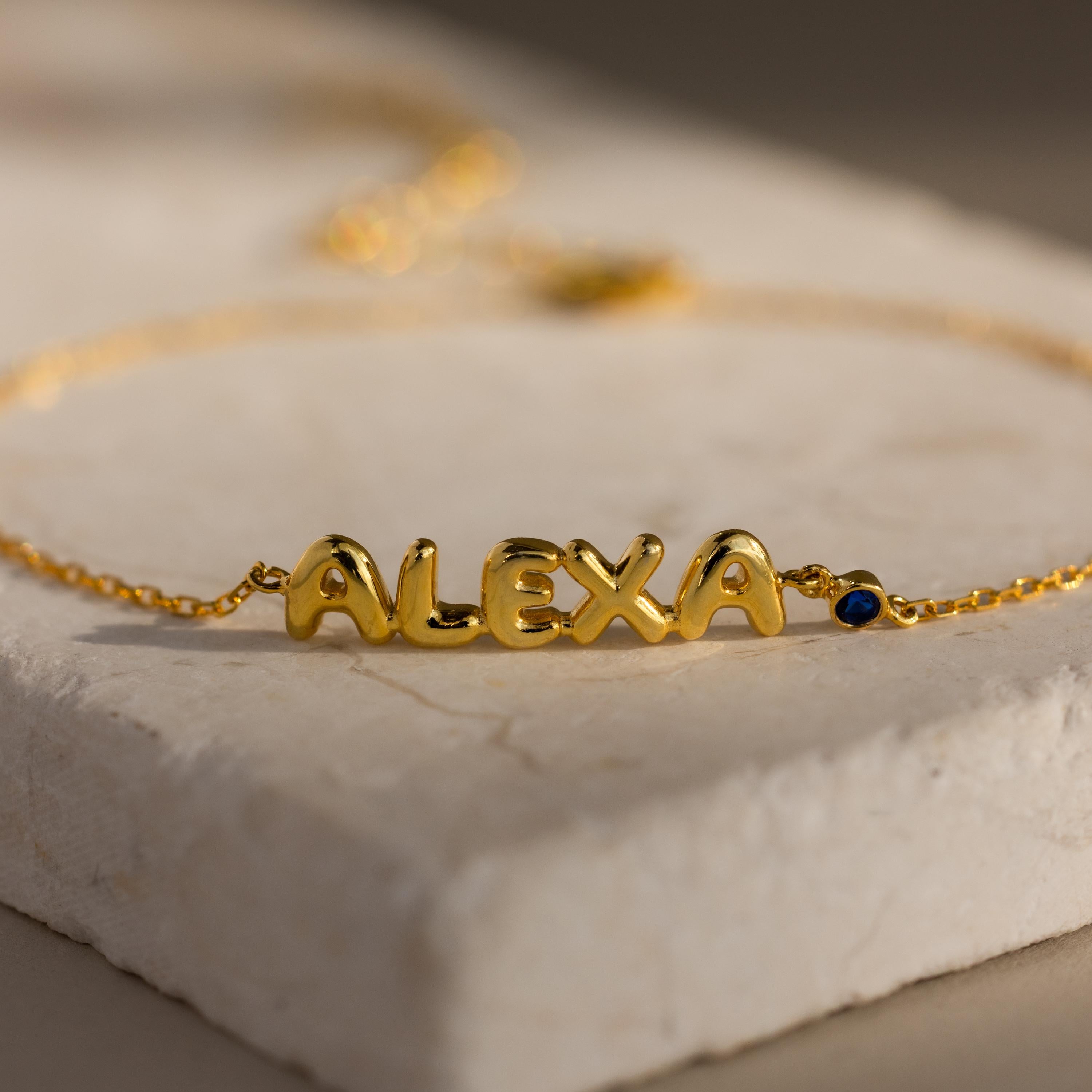 Close-up of the gold “ALEXA” bracelet with a blue birthstone charm displayed on a textured cream surface.