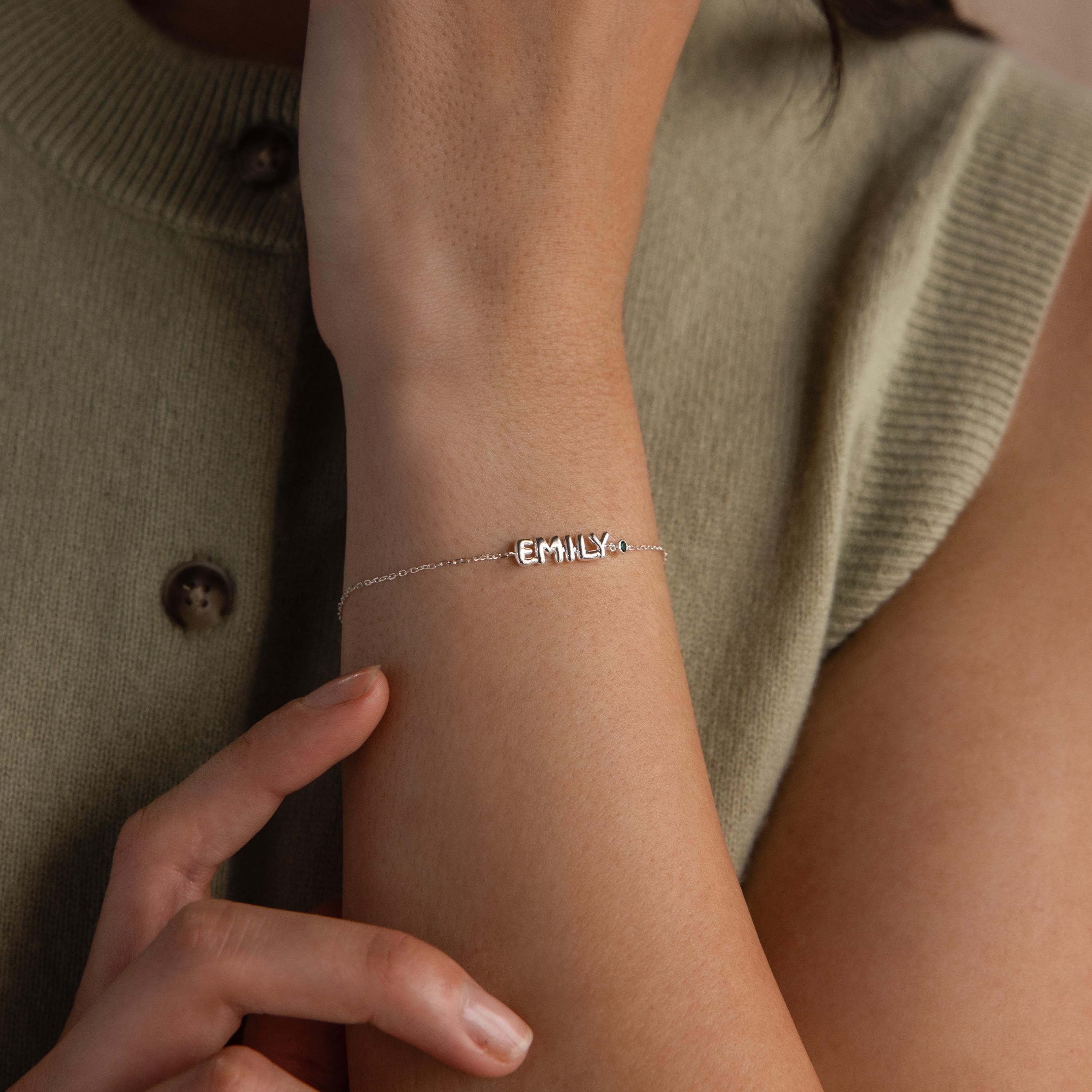 A silver bubble letter name bracelet spelling “EMILY” with a green emerald birthstone is worn on the wrist with an olive-toned sleeveless top.