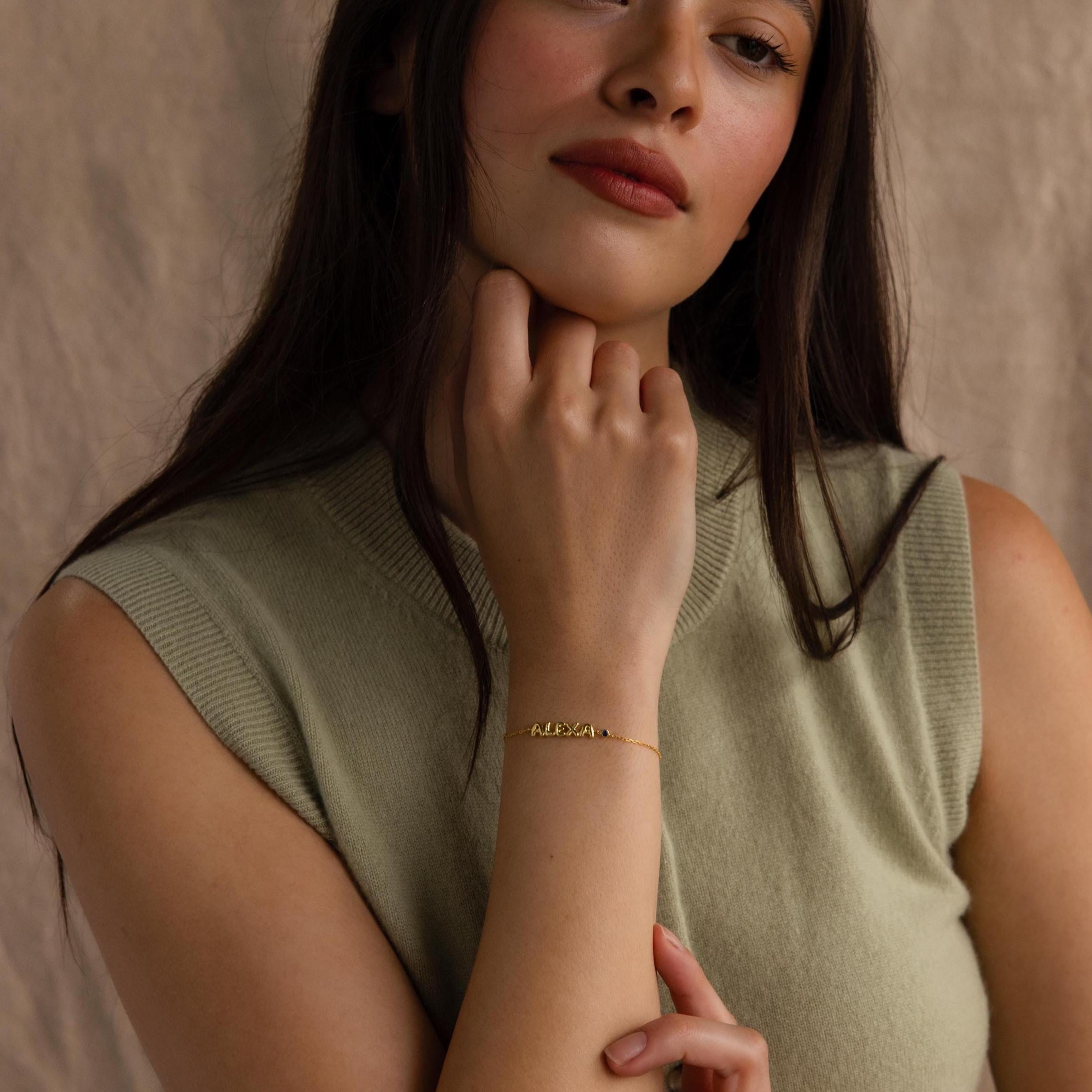 A woman poses wearing the gold “ALEXA” bracelet while resting her chin on her hand.