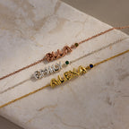 Three name bracelets—“ELLA” in rose gold, “EMILY” in silver, and “ALEXA” in gold—each paired with a birthstone charm, are displayed on a marble surface.