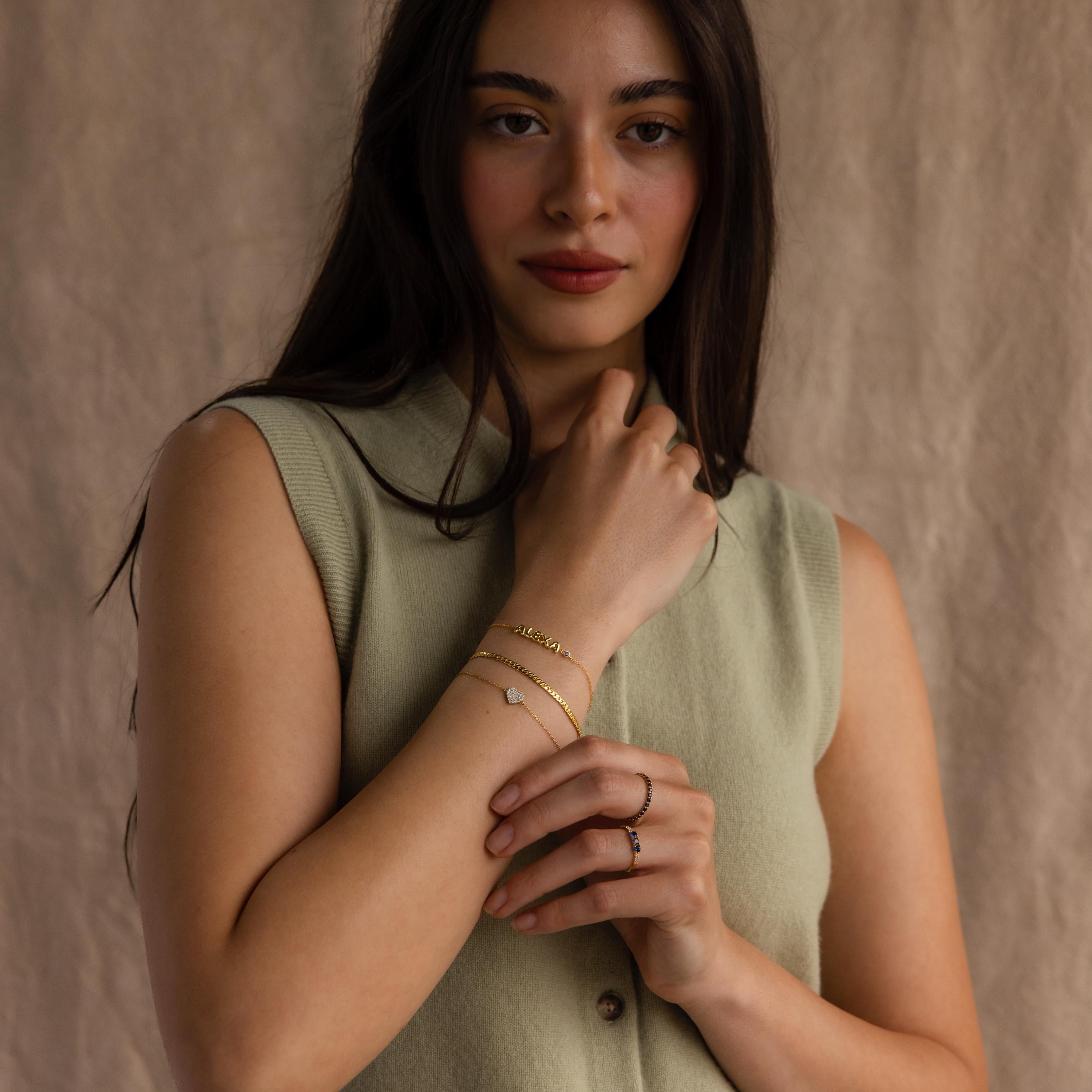 Far view of model wearing a dainty gold bubble letter bracelet with the name "ALEXA", stacked with a snake chain bracelet and pave heart bracelet.