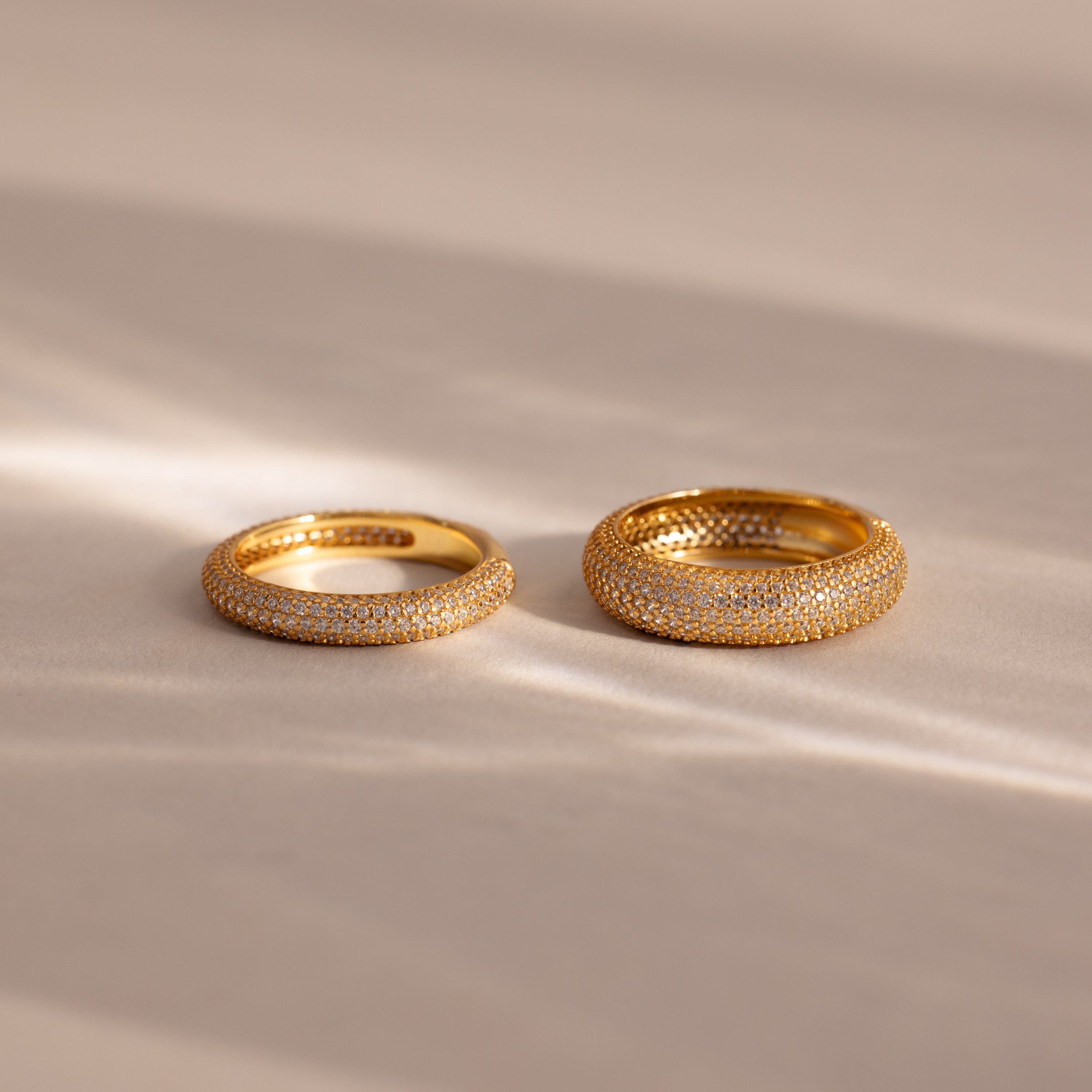Two Pave Stacking Rings adorned with small, sparkling stones are shown on a soft beige surface under gentle lighting.