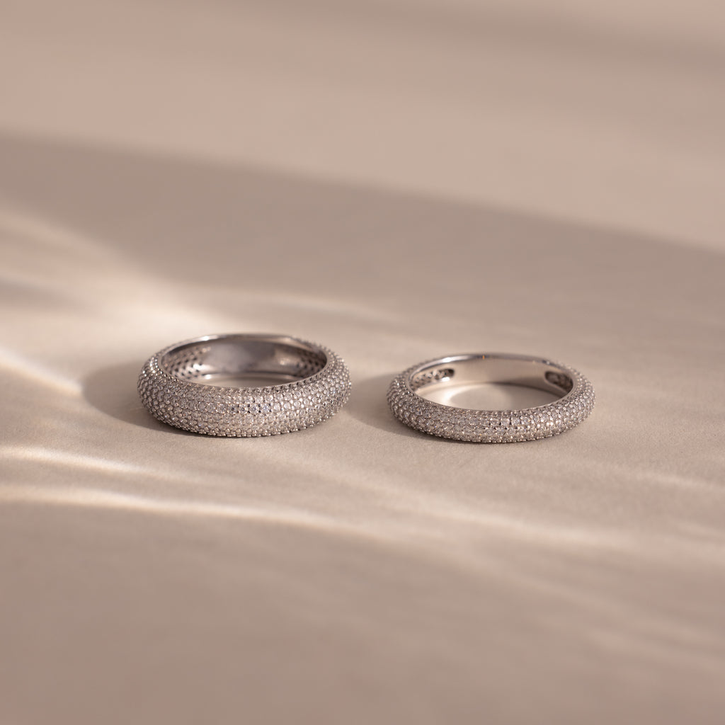 Two Pave Stacking Rings in silver, adorned with diamond-studded details, rest on a soft beige surface as gentle light and shadows accentuate their sparkling bands.