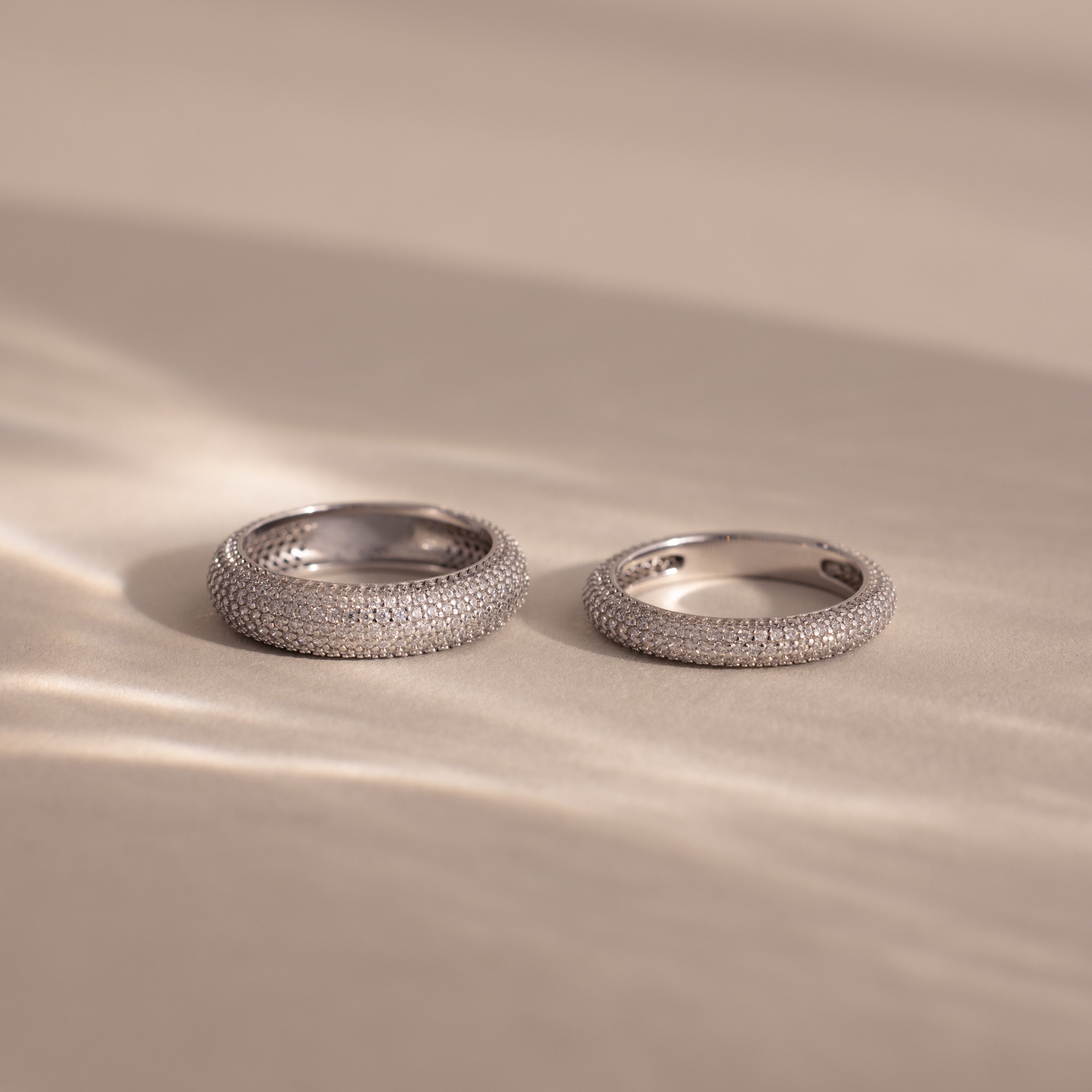 Two Pave Stacking Rings in silver, adorned with diamond-studded details, rest on a soft beige surface as gentle light and shadows accentuate their sparkling bands.