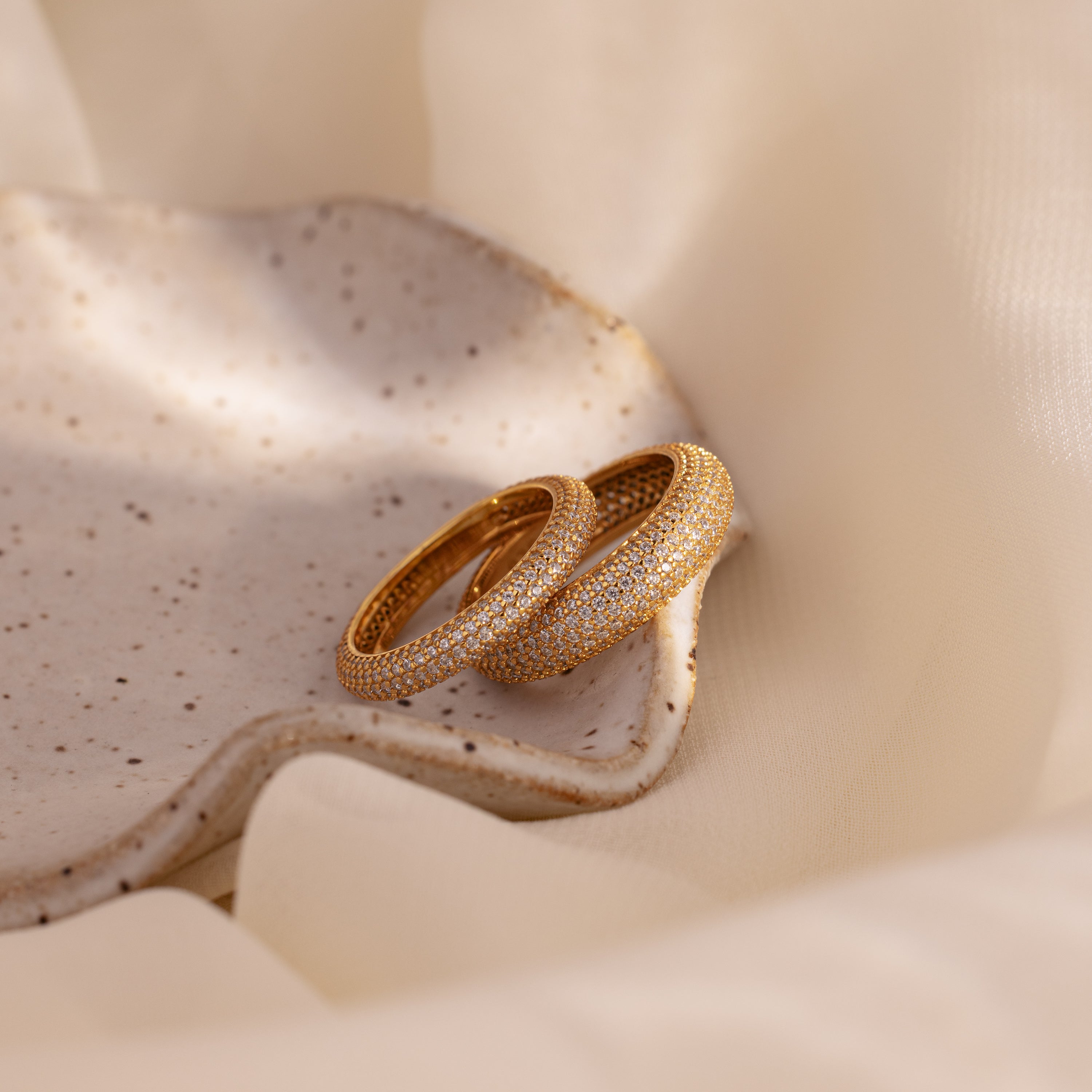 Two Pave Stacking Rings with shimmering diamonds are displayed on a speckled ceramic dish, accented by soft beige fabric.