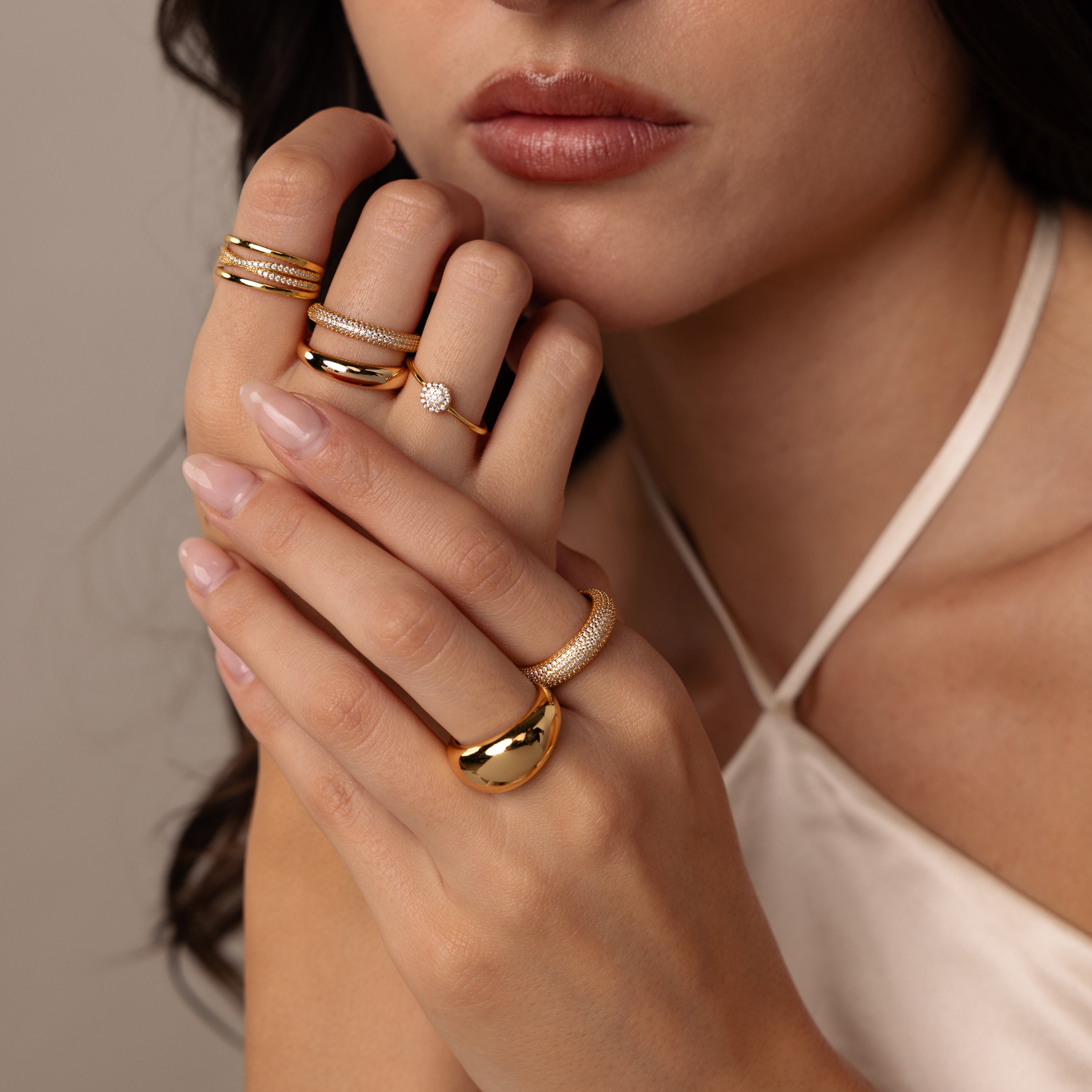 A woman with manicured nails wears multiple gold rings, including Pave Stacking Rings, and a dome band on her fingers while dressed in a cream halter top.