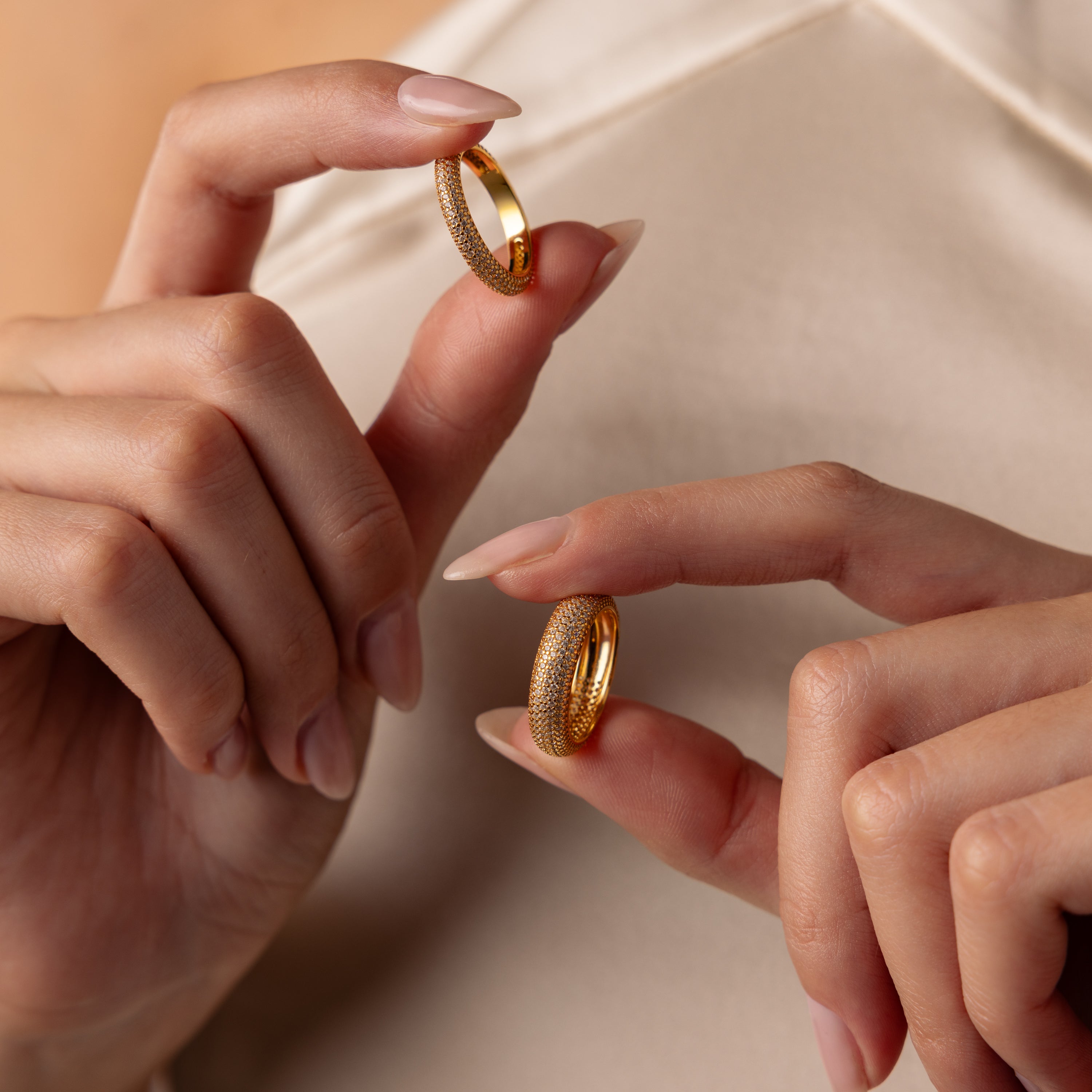Close-up of hands holding two Pave Stacking Rings—a classic wedding band and a dome band—against a cream-colored fabric background.