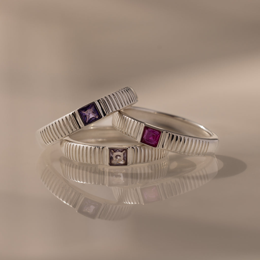 The Ribbed Alexandrite Band, a silver ring with a square-cut purple stone, is stacked with similar rings and reflected on a smooth surface.