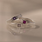 The Ribbed Alexandrite Band, a silver ring with a square-cut purple stone, is stacked with similar rings and reflected on a smooth surface.