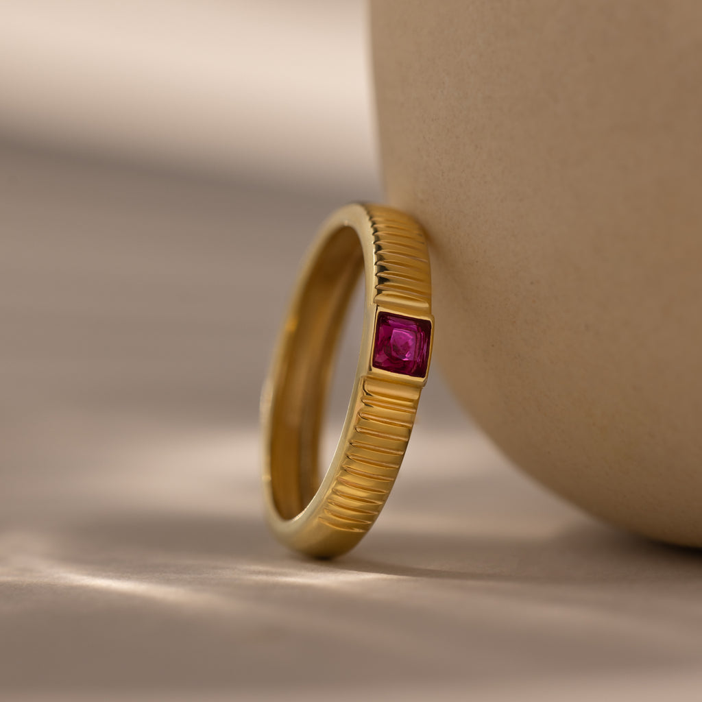The Ribbed Ruby Band, a gold ring with a ribbed band and small square-cut pink stone, leans against a smooth beige surface.