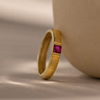 The Ribbed Ruby Band, a gold ring with a ribbed band and small square-cut pink stone, leans against a smooth beige surface.