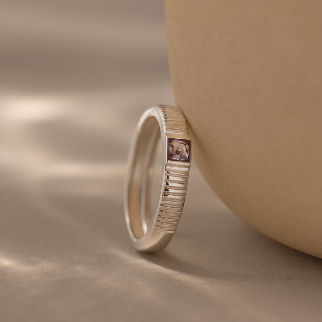 The Ribbed Alexandrite Band, featuring a rectangular purple square-cut stone and a silver ribbed design, is displayed on a smooth beige surface under soft lighting.