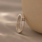 The Ribbed Alexandrite Band, featuring a rectangular purple square-cut stone and a silver ribbed design, is displayed on a smooth beige surface under soft lighting.