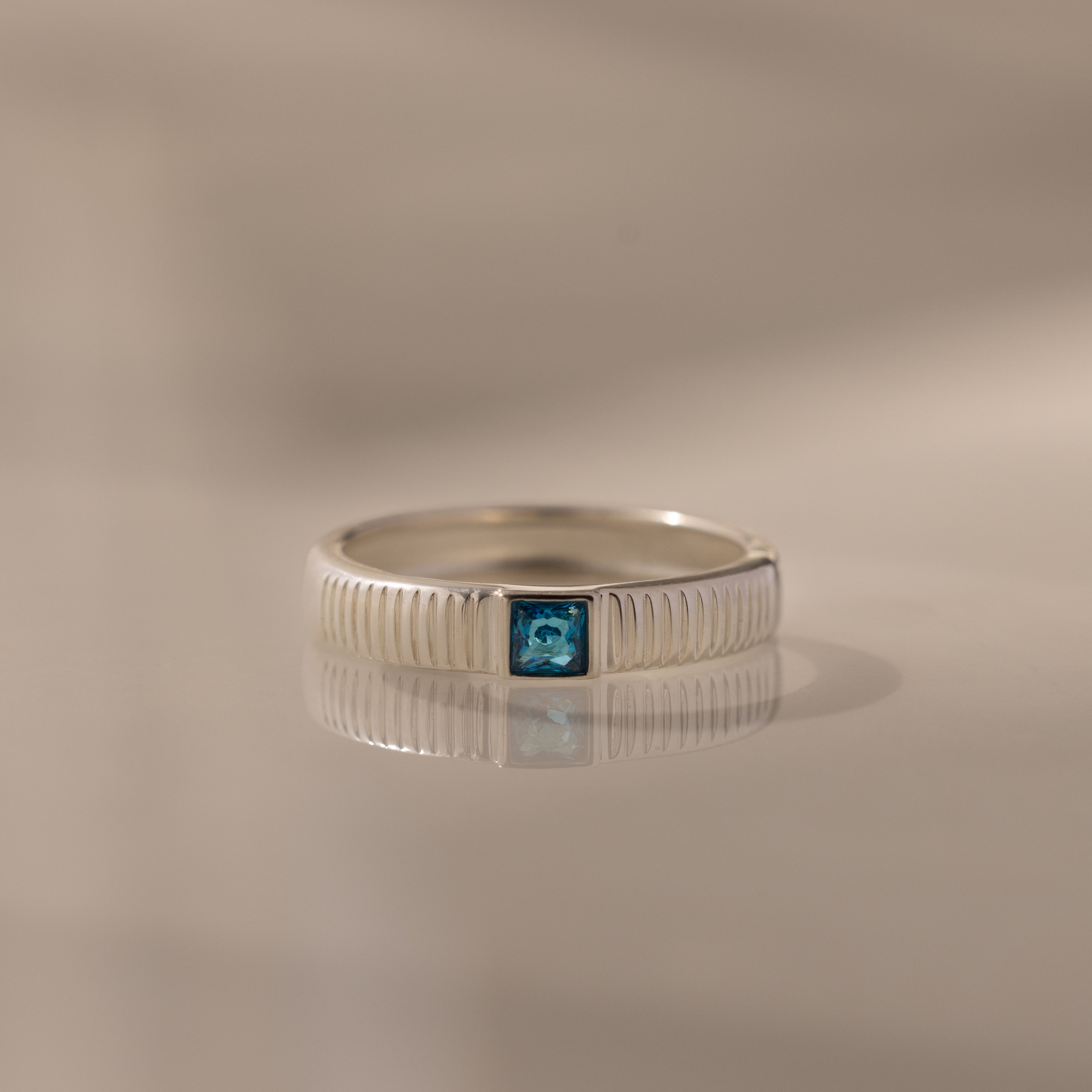 The Ribbed Blue Zircon Band is a silver stacking ring with a central blue square gemstone, shown on a reflective surface.
