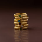 A stack of six gold rings, featuring the Ribbed Peridot Band and square-cut birthstone ring, each set with a unique colored gemstone, displayed against a brown background.