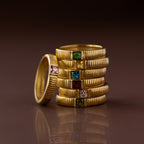 The Ribbed Garnet Band is stacked with gold rings featuring various colored gemstones, all neatly arranged on a reflective brown surface.