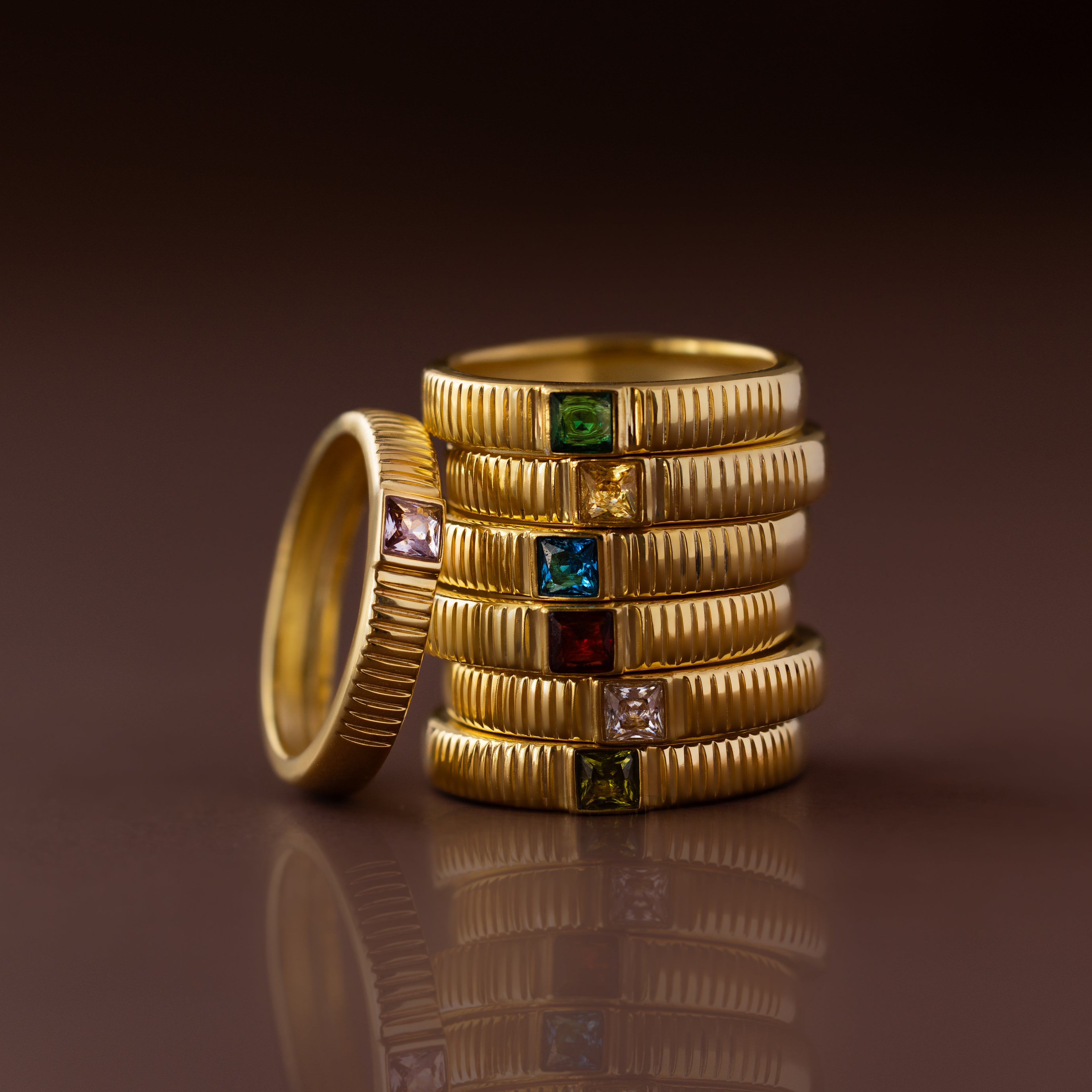 A stack of Ribbed Birthstone Bands, each featuring a different colored gemstone, displayed on a glossy brown surface.