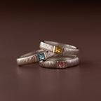 Three silver rings with square blue, yellow, and pink gemstones are stacked on a brown surface—a unique birthstone combination that pairs beautifully with our Ribbed Aquamarine Band.