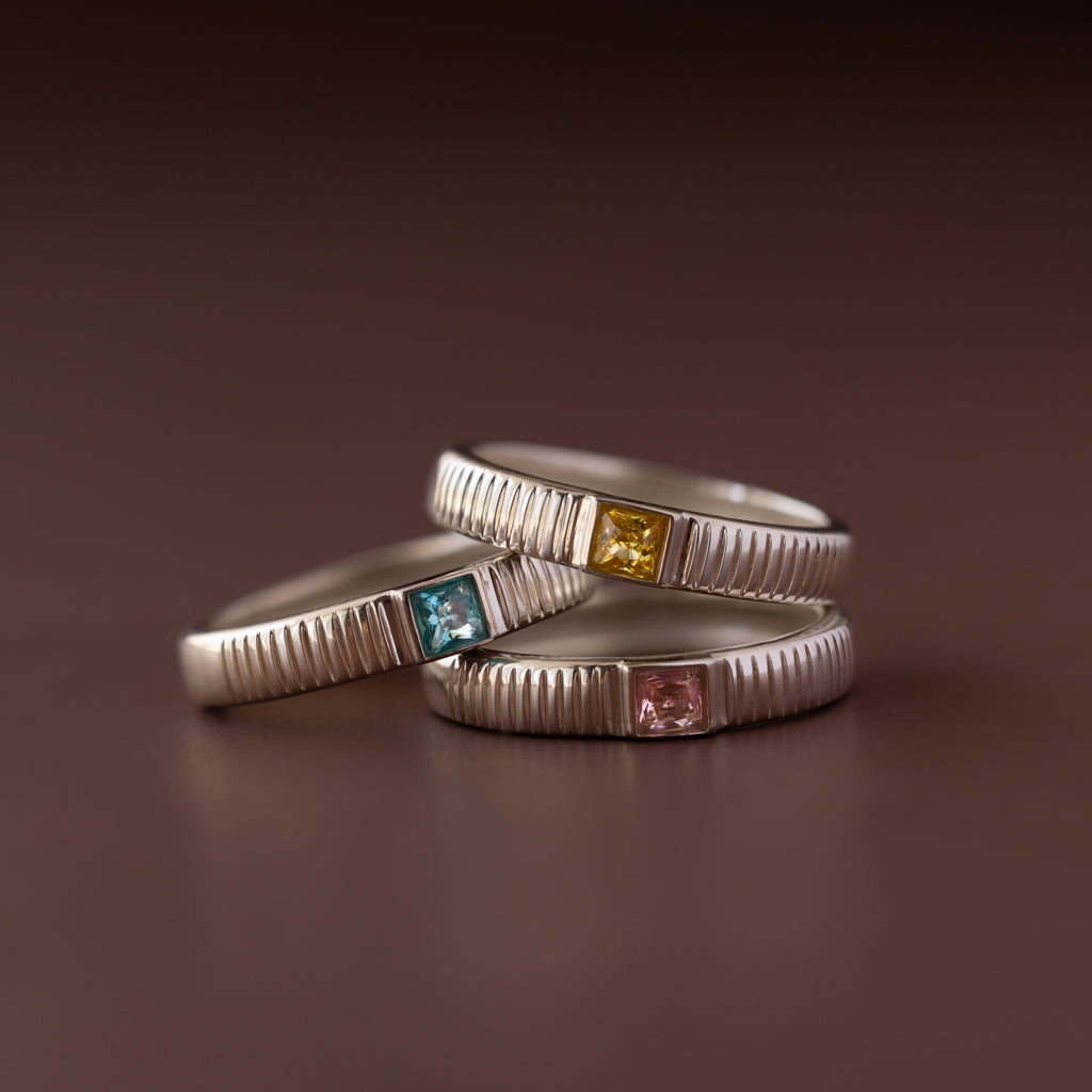 Three silver stacking rings with ribbed bands, each featuring a colored gemstone—blue, yellow, and the Ribbed Pink Tourmaline Band—ideal as unique birthstone rings.