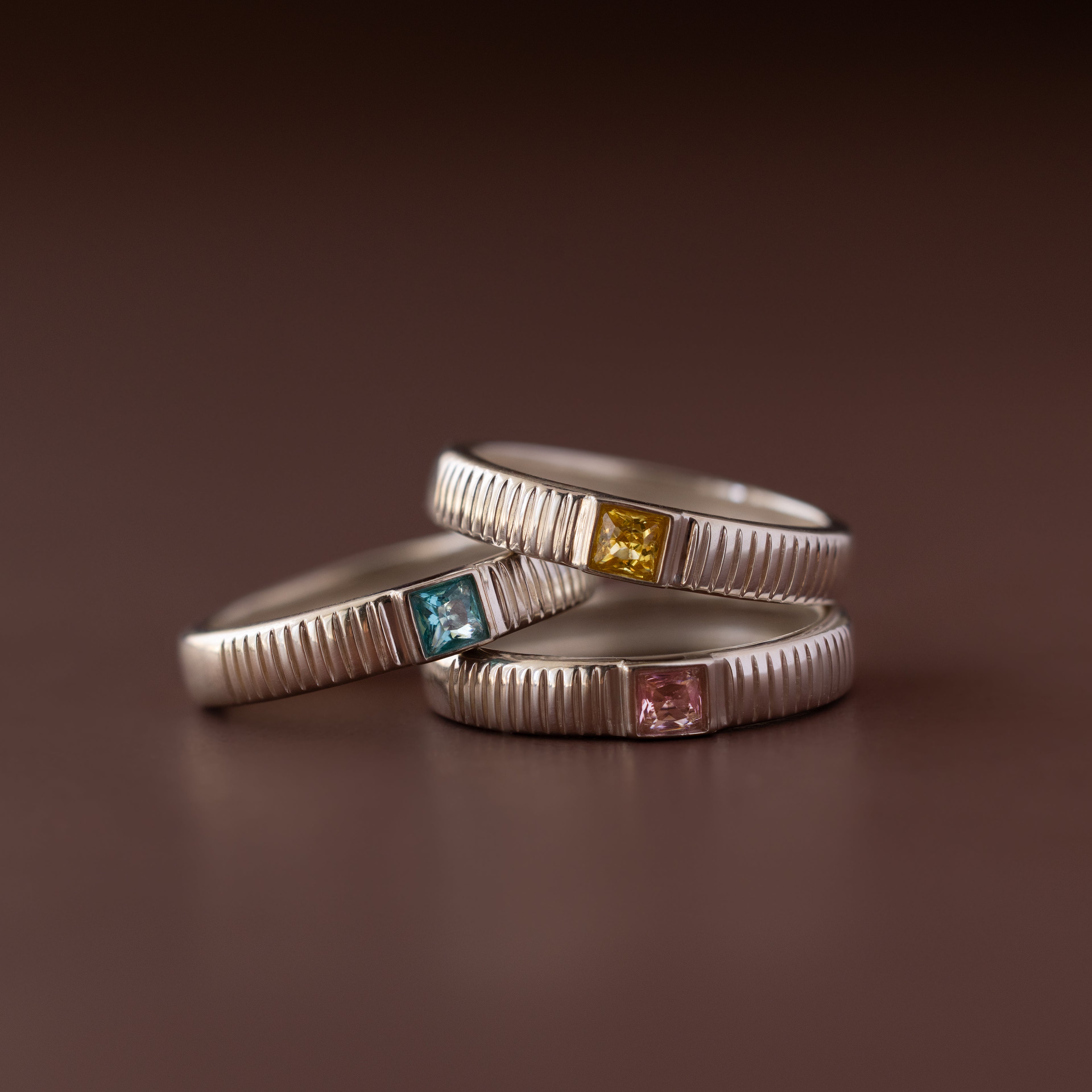 Three silver stacking rings with ribbed bands, each featuring a colored gemstone—blue, yellow, and the Ribbed Pink Tourmaline Band—ideal as unique birthstone rings.