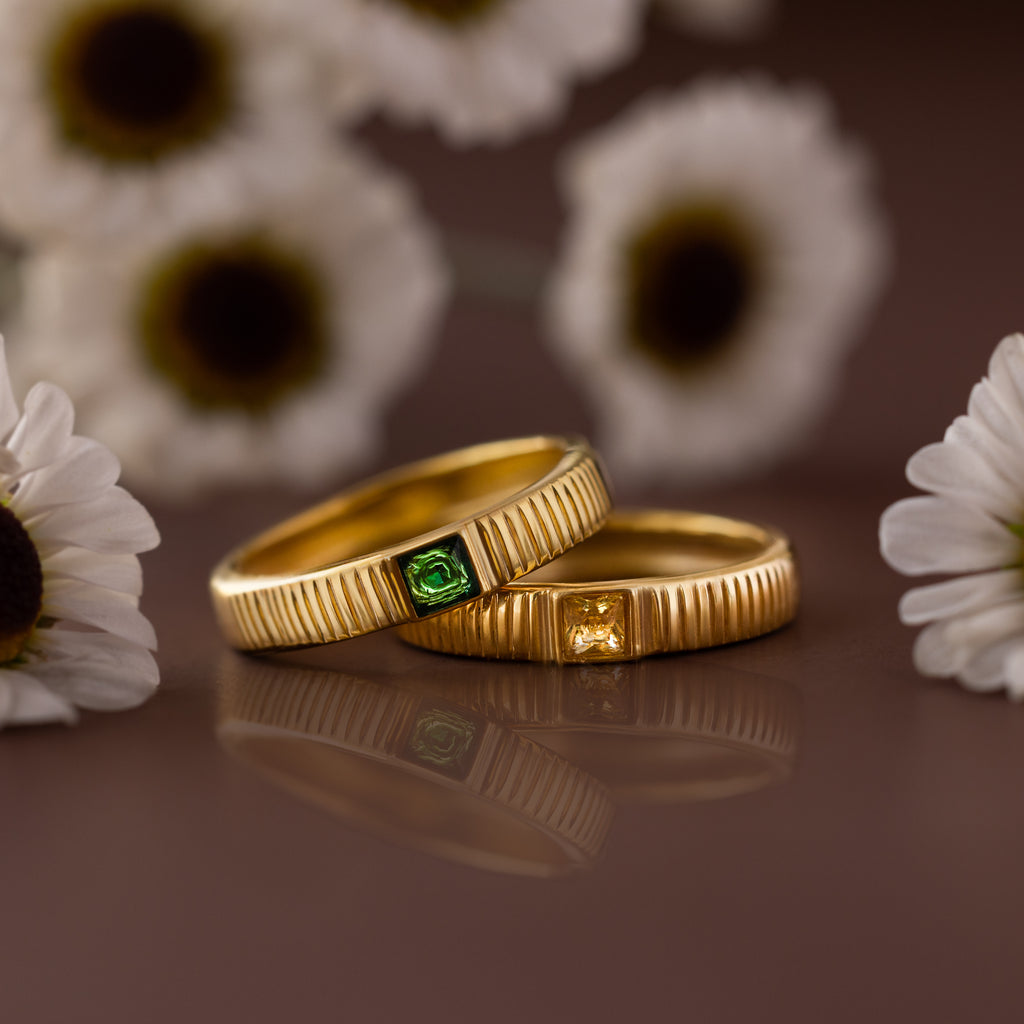 The Ribbed Emerald Band, a gold ring with a green square-cut stone, is displayed among white daisies beside another yellow-stoned ring, creating an eye-catching birthstone jewelry display.