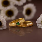 The Ribbed Emerald Band, a gold ring with a green square-cut stone, is displayed among white daisies beside another yellow-stoned ring, creating an eye-catching birthstone jewelry display.