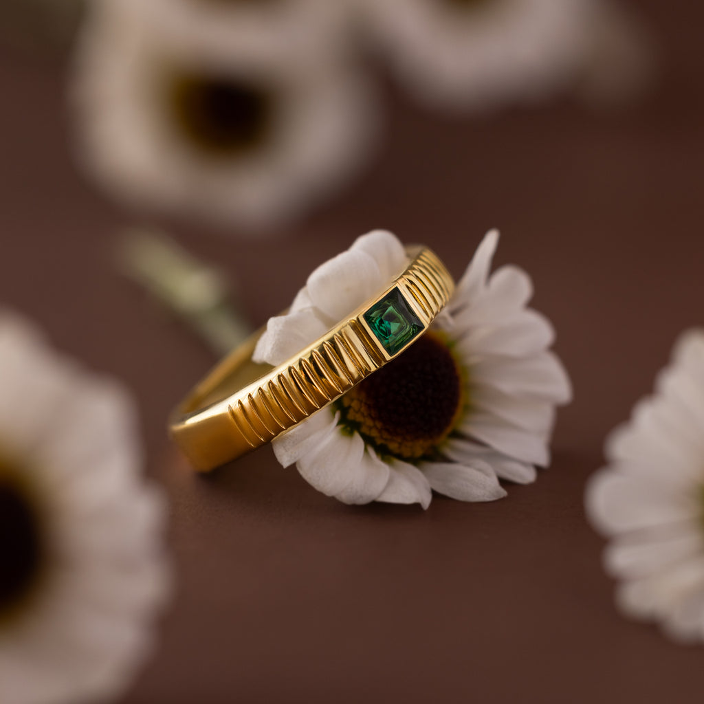 The Ribbed Emerald Band with a square-cut green gemstone rests atop a white daisy, with more daisies softly blurred in the background.