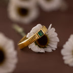 The Ribbed Emerald Band with a square-cut green gemstone rests atop a white daisy, with more daisies softly blurred in the background.
